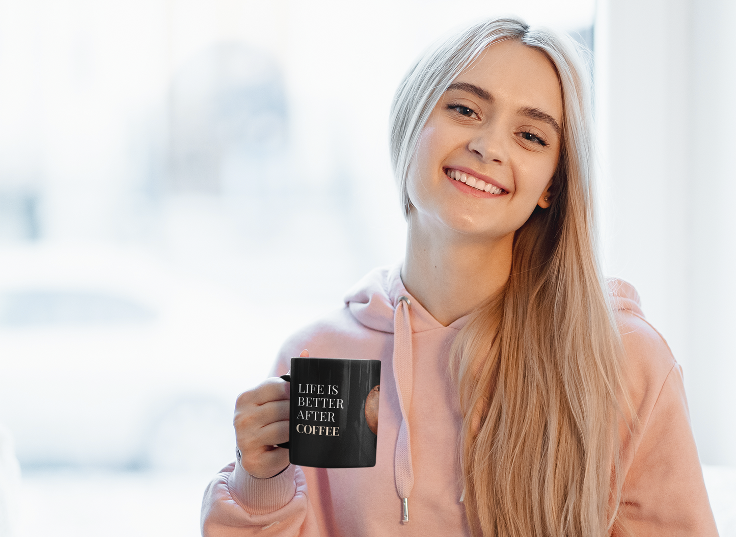 A smiling young woman with long blonde hair holds the "Life Is Better After Coffee" Black Mug (350 ML)—perfect for daily use or as a gift. She wears a pink hoodie and sits in a bright, softly lit room.
